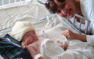 A woman is leaning over a hospital bed with a baby lying in it. The baby has a soft cap on their head with medical tubes and wires attached, indicating they are receiving medical care. The woman is smiling warmly and looking at the camera, while the baby appears to be asleep or resting. The setting is clearly a medical facility, as evidenced by the equipment and the hospital bed.