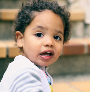 A young child with curly hair, wearing a light-colored shirt with striped sleeves, looks directly at the camera with a curious expression. The background features blurred steps and bricks, suggesting the child is outdoors.