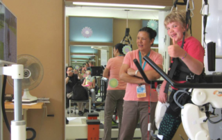 A child wearing a red shirt and black pants is standing with the assistance of a robotic exoskeleton device in a therapy session. The device is labeled "Hocoma." A woman, likely a therapist, is standing next to the child, smiling and interacting with them. In the background, there are mirrors reflecting the scene, showing multiple angles of the child and the therapist. A person is seated and taking a photo or video of the session. There are flower decorations on the walls, and a fan and some papers are on a small table near a monitor displaying an image.