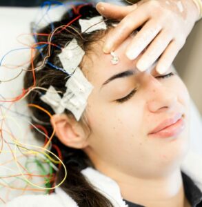 Young female undergoing an electroencephalogram (EEG) test. The person has multiple electrodes attached to their scalp with adhesive pads, and colorful wires are connected to the electrodes. The individual appears calm, with eyes closed and a slight smile on their face, as a hand gently adjusts one of the electrodes on their forehead.