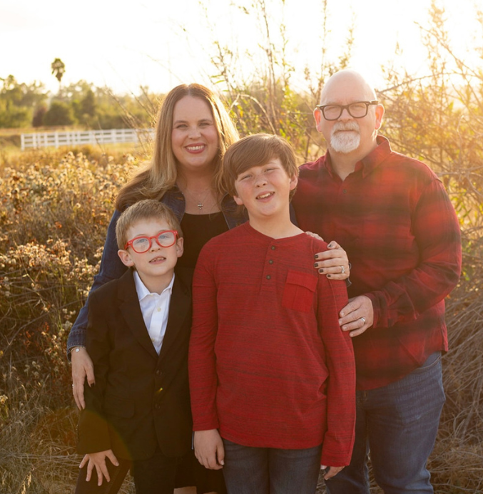 This is a family photo taken outdoors in natural sunlight. The family consists of two adults and two children, all standing close together and smiling. The mother, with long blonde hair, is standing next to the younger child, who is wearing a black suit and red glasses. The father, with a bald head, white beard, and glasses, is wearing a red plaid shirt and standing next to the older child, who is wearing a red long-sleeve shirt. The background is a sunny, rustic field with some plants and trees, and there is a white fence visible in the distance. The sun is shining behind the family, casting a warm, golden glow over the scene.