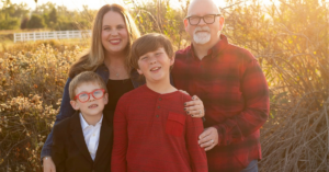 This is a family photo taken outdoors in natural sunlight. The family consists of two adults and two children, all standing close together and smiling. The mother, with long blonde hair, is standing next to the younger child, who is wearing a black suit and red glasses. The father, with a bald head, white beard, and glasses, is wearing a red plaid shirt and standing next to the older child, who is wearing a red long-sleeve shirt. The background is a sunny, rustic field with some plants and trees, and there is a white fence visible in the distance. The sun is shining behind the family, casting a warm, golden glow over the scene.