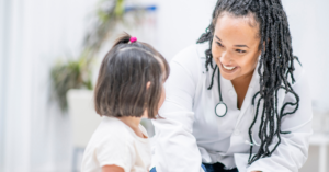 This image shows a doctor, smiling warmly, interacting with a young child. The doctor, who has long braided hair, is wearing a white coat and a stethoscope, indicating a medical setting. The child, with short hair, is looking at the doctor. The setting appears to be a bright, friendly medical environment, possibly a clinic or doctor's office. The image conveys a sense of care, trust, and positive communication between the healthcare professional and the child.