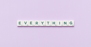 The image shows the word "EVERYTHING" spelled out using Scrabble-like letter tiles. Each tile features a letter with a corresponding point value. The tiles are arranged in a straight line on a soft lavender background.