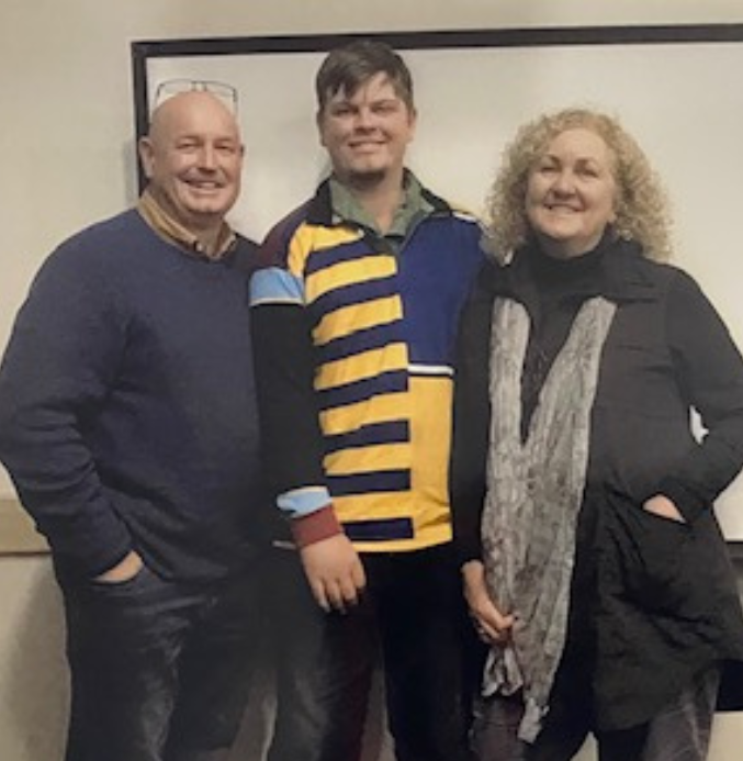 The image shows three adults standing side by side, smiling for the camera. On the left is a man with a shaved head wearing a dark blue sweater and jeans. In the middle is a younger man wearing a colorful rugby-style shirt with bold horizontal stripes in blue, yellow, and black, along with dark pants. On the right is a woman with curly blonde hair, wearing a black jacket and a gray scarf. All three have their hands in their pockets or resting comfortably. The background features a plain whiteboard, suggesting an indoor setting, possibly a classroom or meeting room. The mood of the image feels friendly and casual.