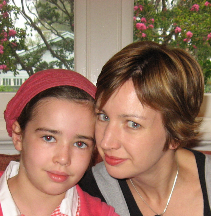 The image shows two people sitting close together, both looking directly at the camera. On the left is a young girl wearing a red headscarf and a pink jacket over a white shirt. On the right is an adult woman with short, light brown hair. They both have light skin, and the woman is slightly leaning in toward the girl, smiling gently. Behind them, there is a window with vibrant greenery and pink flowers visible through the glass. The setting feels warm and personal, as if taken in a cozy home environment.