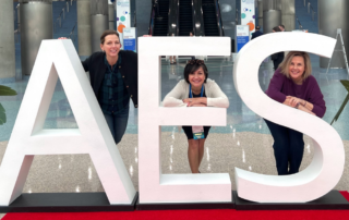 Three women are posing in front of large white letters that spell "AES" at an indoor event space. Each woman is leaning playfully on or around one of the letters. The woman on the left, wearing a black jacket and plaid shirt, is leaning on the "A." The woman in the center, dressed in a white sweater and black skirt, leans through the "E" with her arms resting on it. The woman on the right, wearing a purple sweater, poses with her arms leaning on the "S." The space features large concrete columns, escalators in the background, and potted plants on either side of the display. A red carpet lies beneath the letters.