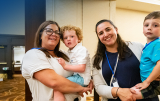 Two mothers holding their young boys. The mothers are smiling and they are at a conference.