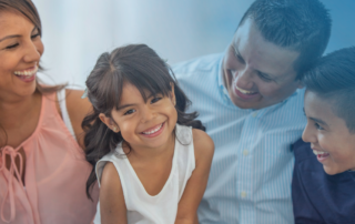 A joyful Hispanic family of four sits close together, smiling and laughing. A young girl in a white dress is in the center, grinning brightly at the camera. Her mother, wearing a sleeveless pink blouse, is on the left; her father, in a light blue striped shirt, and her older brother, in a dark navy shirt, are on the right. The background is softly blurred with a blue gradient, emphasizing the family's warmth and happiness.