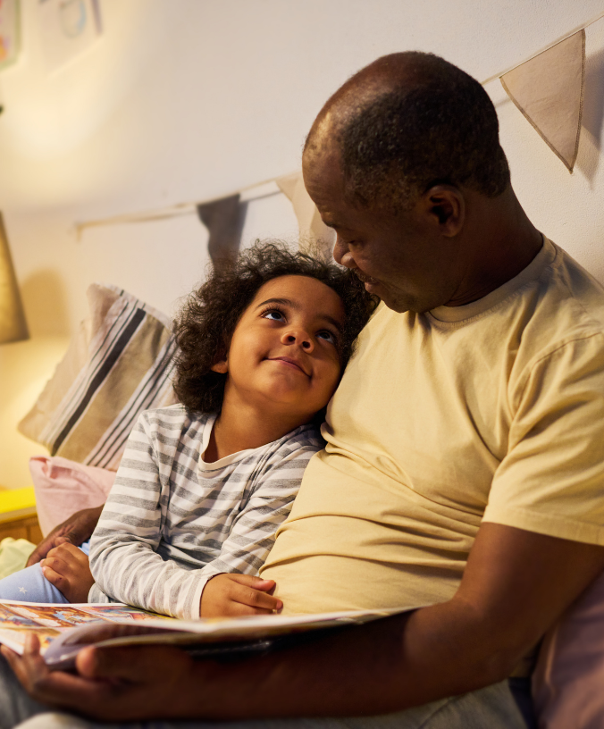 A father and young girl are sitting on a bed. He is reading to her and she is smiling at him.