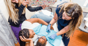A child lies on a medical exam table with a caregiver beside them, gently comforting them. A medical professional in gloves prepares to apply a bandage or conduct a procedure on the child’s abdomen. Another staff member holds up a colorful distraction board covered in stickers, helping keep the child calm and engaged.