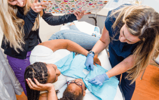 A child lies on a medical exam table with a caregiver beside them, gently comforting them. A medical professional in gloves prepares to apply a bandage or conduct a procedure on the child’s abdomen. Another staff member holds up a colorful distraction board covered in stickers, helping keep the child calm and engaged.
