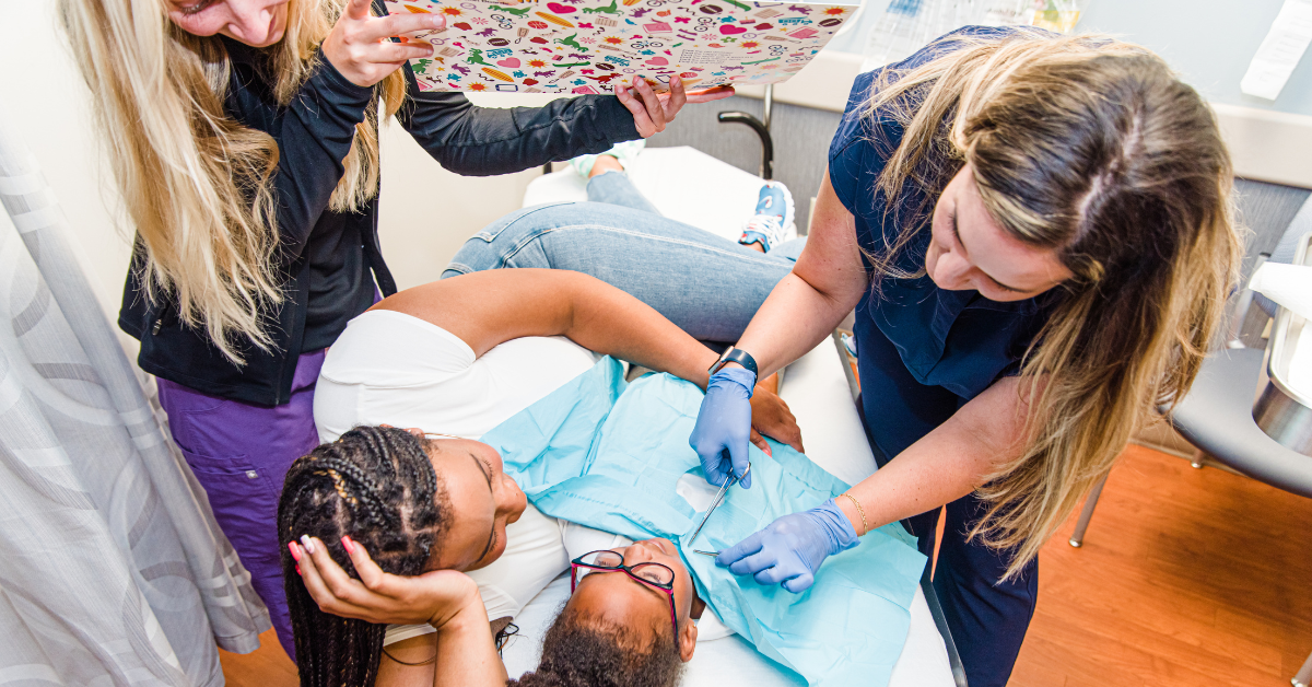 A child lies on a medical exam table with a caregiver beside them, gently comforting them. A medical professional in gloves prepares to apply a bandage or conduct a procedure on the child’s abdomen. Another staff member holds up a colorful distraction board covered in stickers, helping keep the child calm and engaged.