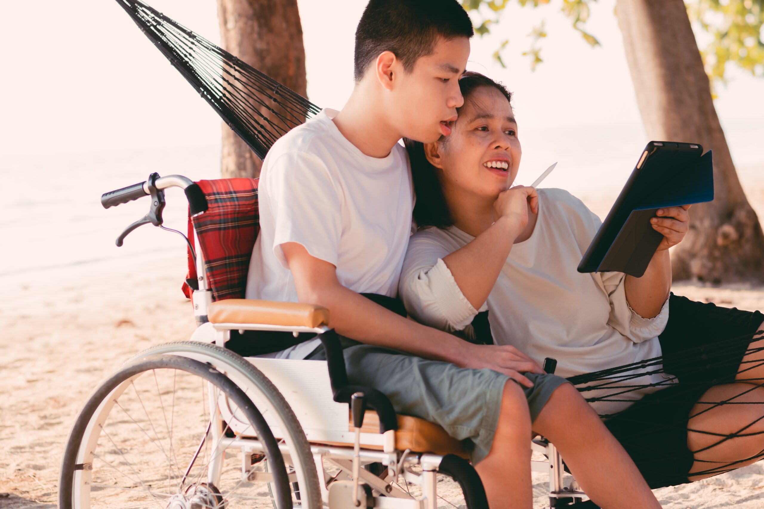 An adult sibling lays in a hammock and reads from a tablet to her brother who is seated next to her in a wheelchair.