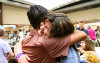 Two people hugging tightly in a conference hall, the woman smiling with her eyes closed; tables, chairs, and balloons in the background.