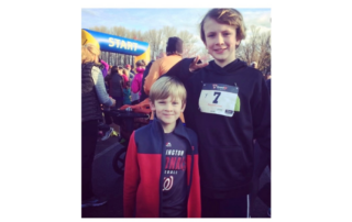 Two boys standing. They are smiling. One is older than the other.