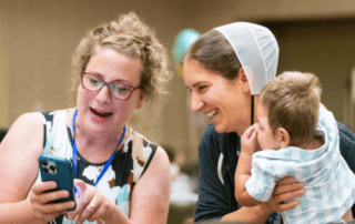 Two women are smiling and looking at a smart phone. One woman is holding a baby.