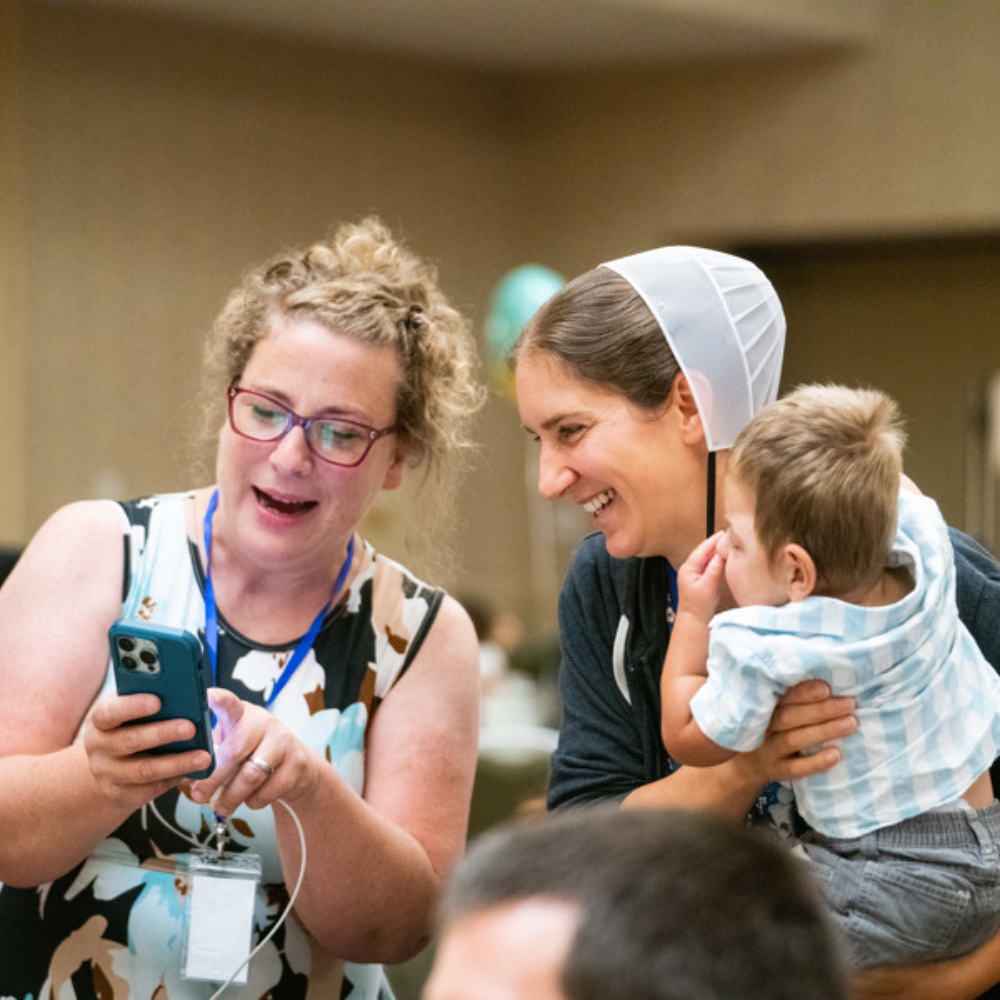 Two women are smiling and looking at a smart phone. One woman is holding a baby.
