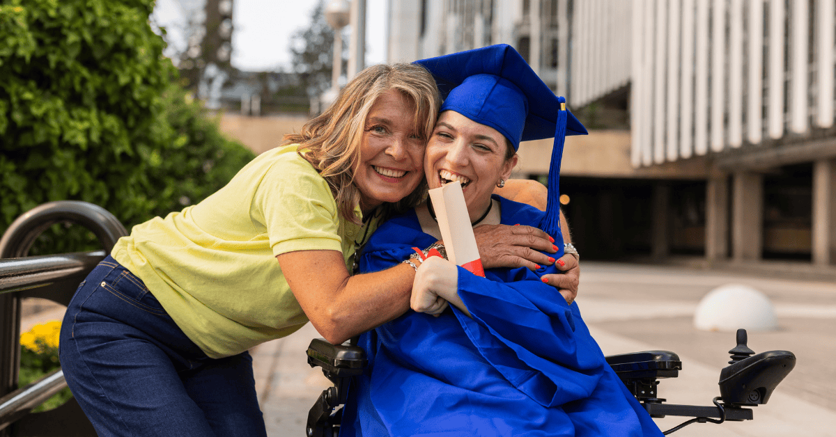 A young woman in a wheelchair and a blue cap and gown holds her diploma while hugging her mom in a yellow shirt and bluejeans.